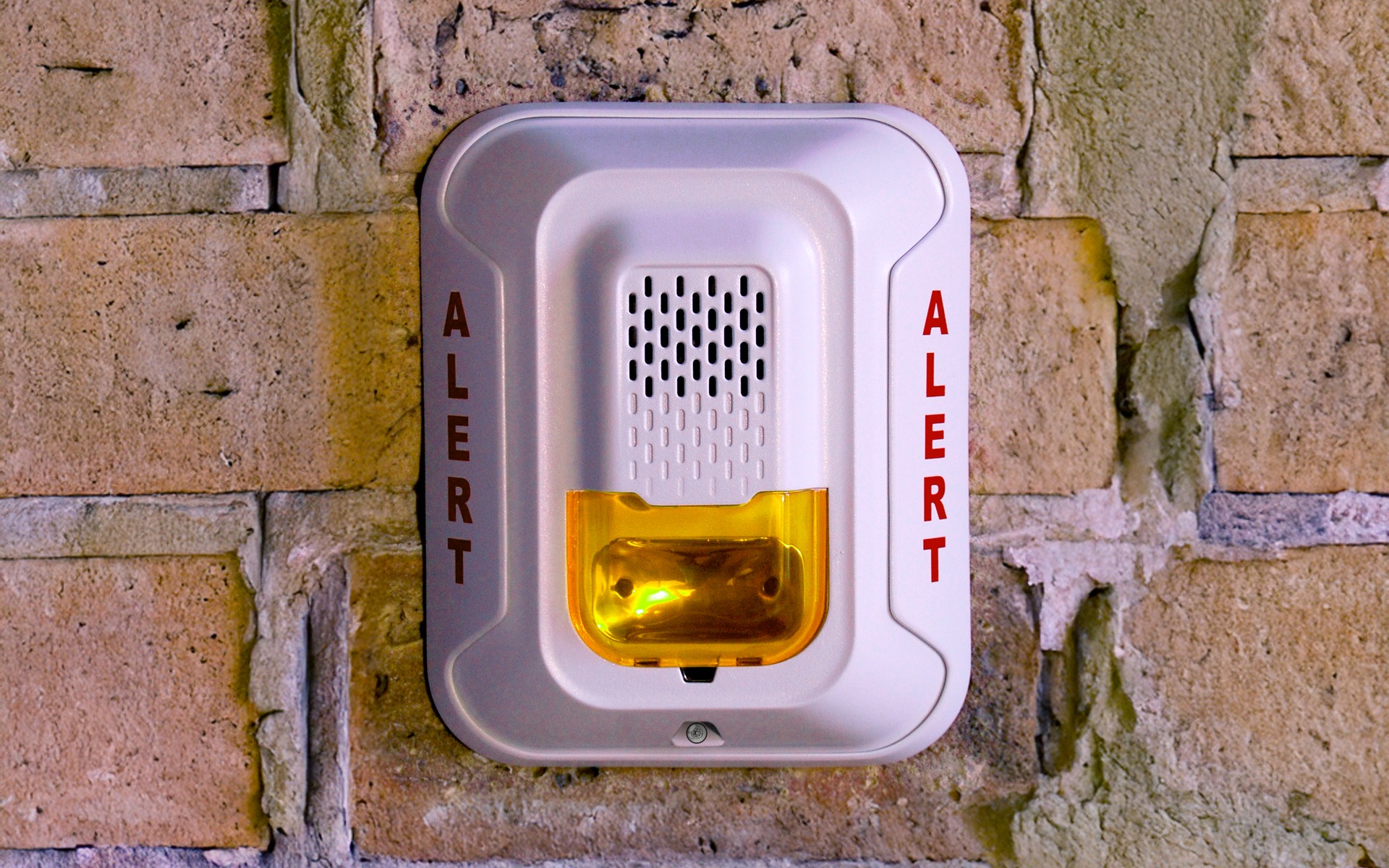 Wall-mounted fire alarm notification appliance with amber strobe lens and ALERT lettering on brick